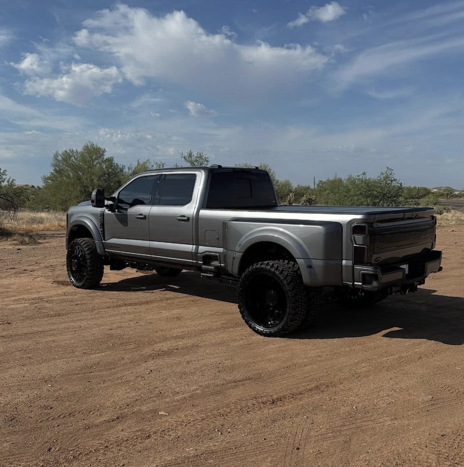 Silver pickup truck with large black wheels on a dirt road under a blue sky.